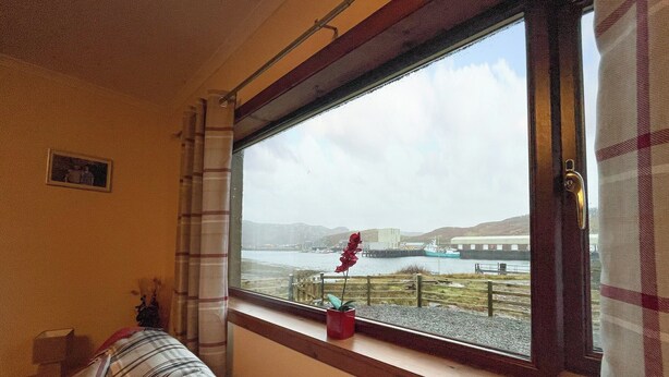 A room with a large window overlooking a body of water and hills. A plaid sofa is positioned near the window, and a small red flower vase is on the windowsill.