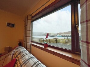 A room with a large window overlooking a body of water and hills. A plaid sofa is positioned near the window, and a small red flower vase is on the windowsill. - 3 bedroom accommodation in Kinlochbervie (Kinlochbervie)