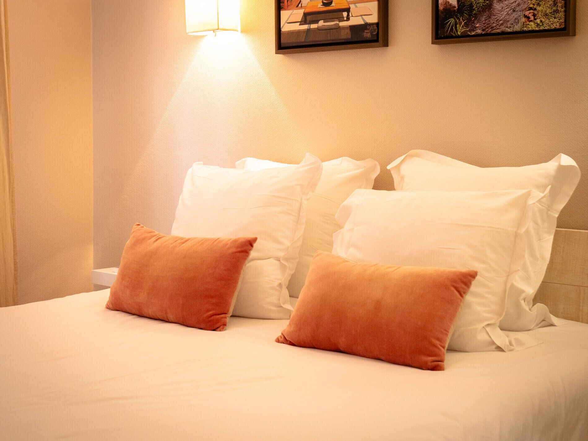 A neatly made bed with white and orange pillows, a wall-mounted picture, and a wall-mounted light.