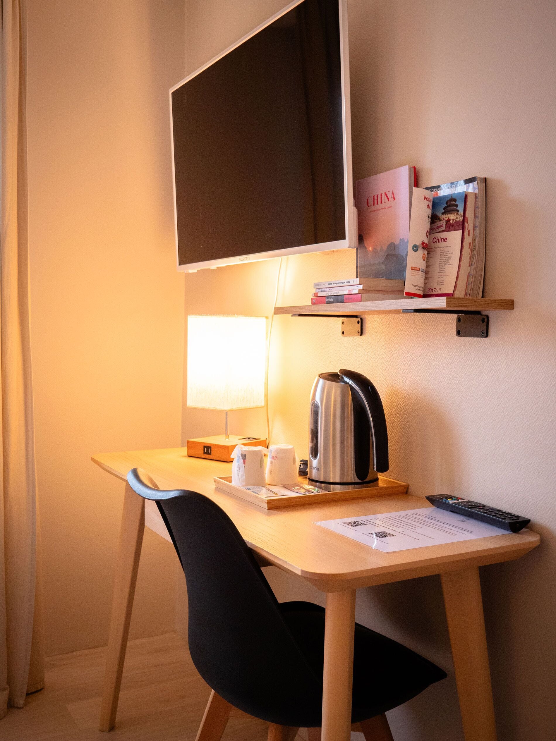 Quarto de hotel com escrivaninha, cadeira, chaleira, comando remoto e televisão na parede.