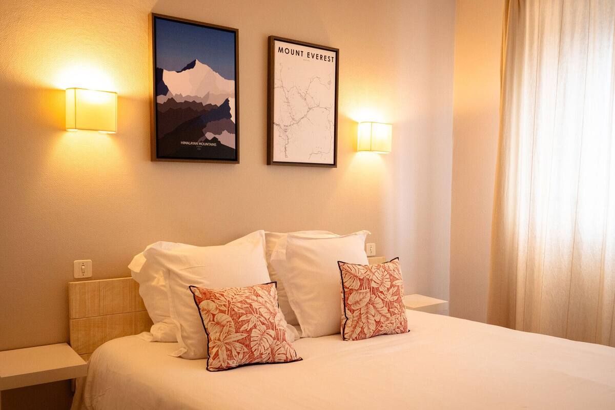 A hotel room with a bed, two wall-mounted lamps, a framed map, and a framed picture of the Himalayas.
