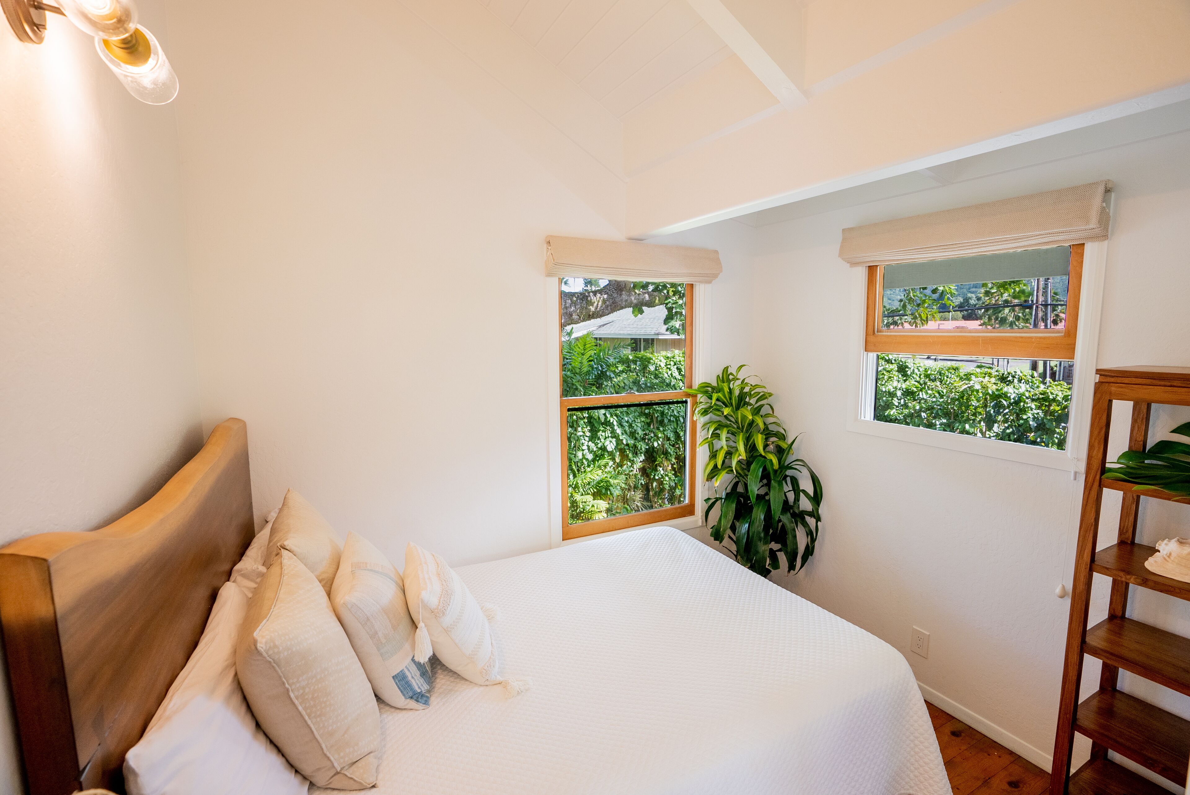 A bedroom with a wooden headboard, a large bed with white bedding, two windows with views of greenery, and a wooden shelf with plants.