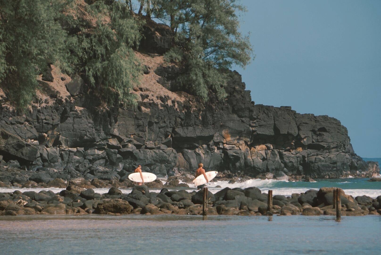 Une côte rocheuse où l’on voit deux surfeurs portant leurs planches de surf.