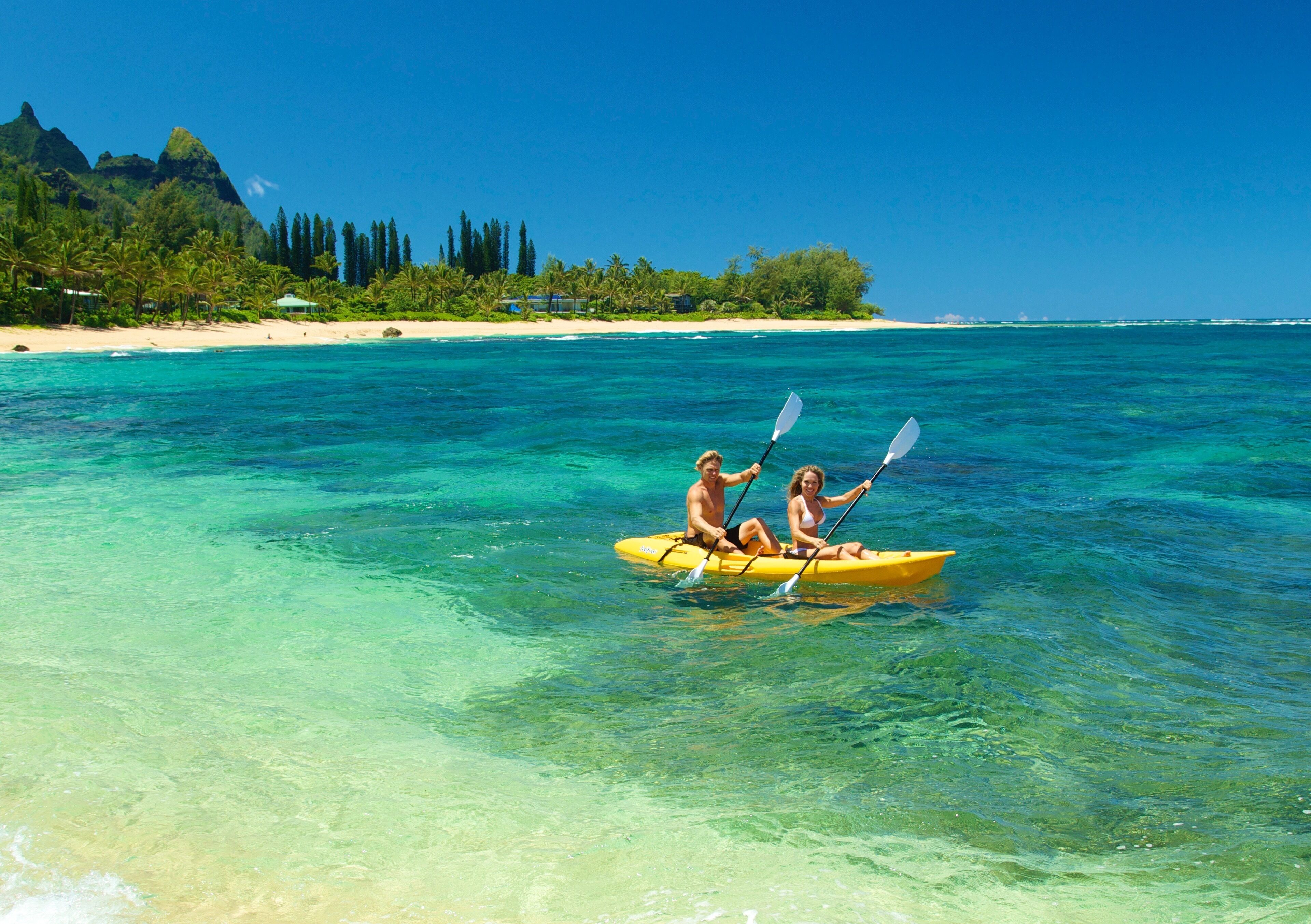 Un couple fait du kayak sur une mer turquoise et limpide, avec une plage et une végétation luxuriante en arrière-plan.