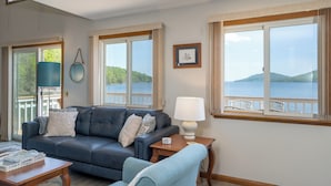 A living room with a sofa, a table, and a lamp. There are large windows with a view of the water and hills. - The Chosen Spot: Secluded, Views, Wineries, Hot tub, Skiing, Pet friendly! (Naples)
