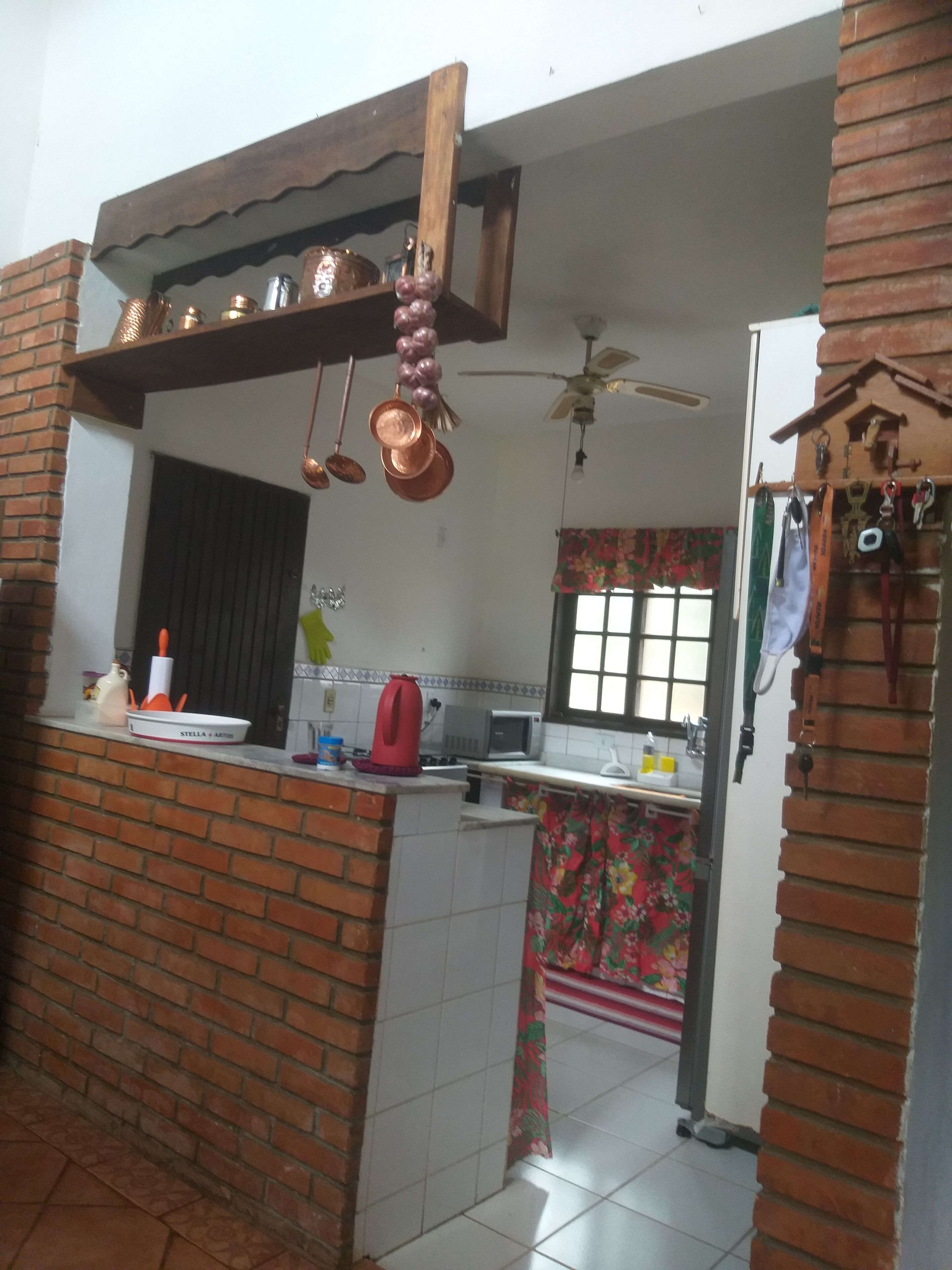 A kitchen with a brick wall, a wooden shelf with hanging pots, a red kettle, and a microwave.