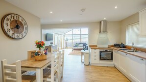 A modern kitchen with a wooden dining table, white chairs, a large clock, and a view through the window. - 2 bedroom accommodation in Llantilio Pertholey (Llantilio Pertholey)