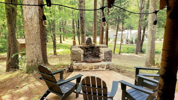 Un espace extérieur aménagé, abrité et doté de chaises Adirondack et d’un foyer en pierre, le tout entouré d’arbres.