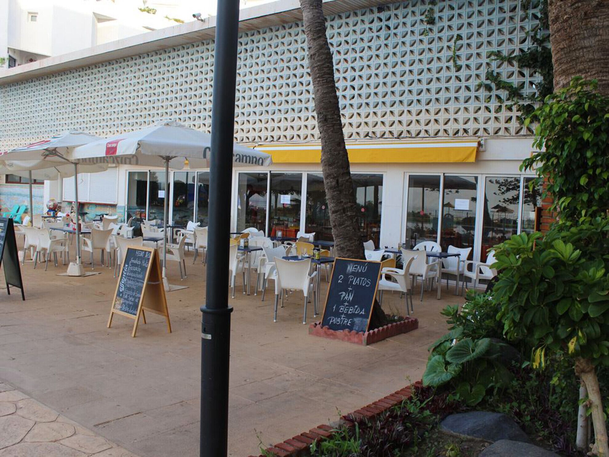An outdoor café with tables and chairs, a chalkboard menu, and a building with a lattice facade in the background.