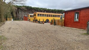Un autobús escolar amarillo estacionado en un camino de grava, con un edificio rojo y una cerca de madera al fondo.