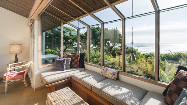 Ein sonnendurchflutetes Zimmer mit einem großen Fenster, das auf einen Garten und das Meer blickt.