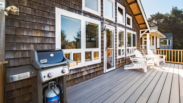 Een houten terras met een barbecue en stoelen, een houten huis met grote ramen, en een heldere lucht.