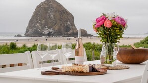 A table set with wine glasses, a bottle of champagne, a wooden board with cheese, and a vase of flowers, overlooking a beach with a large rock formation.