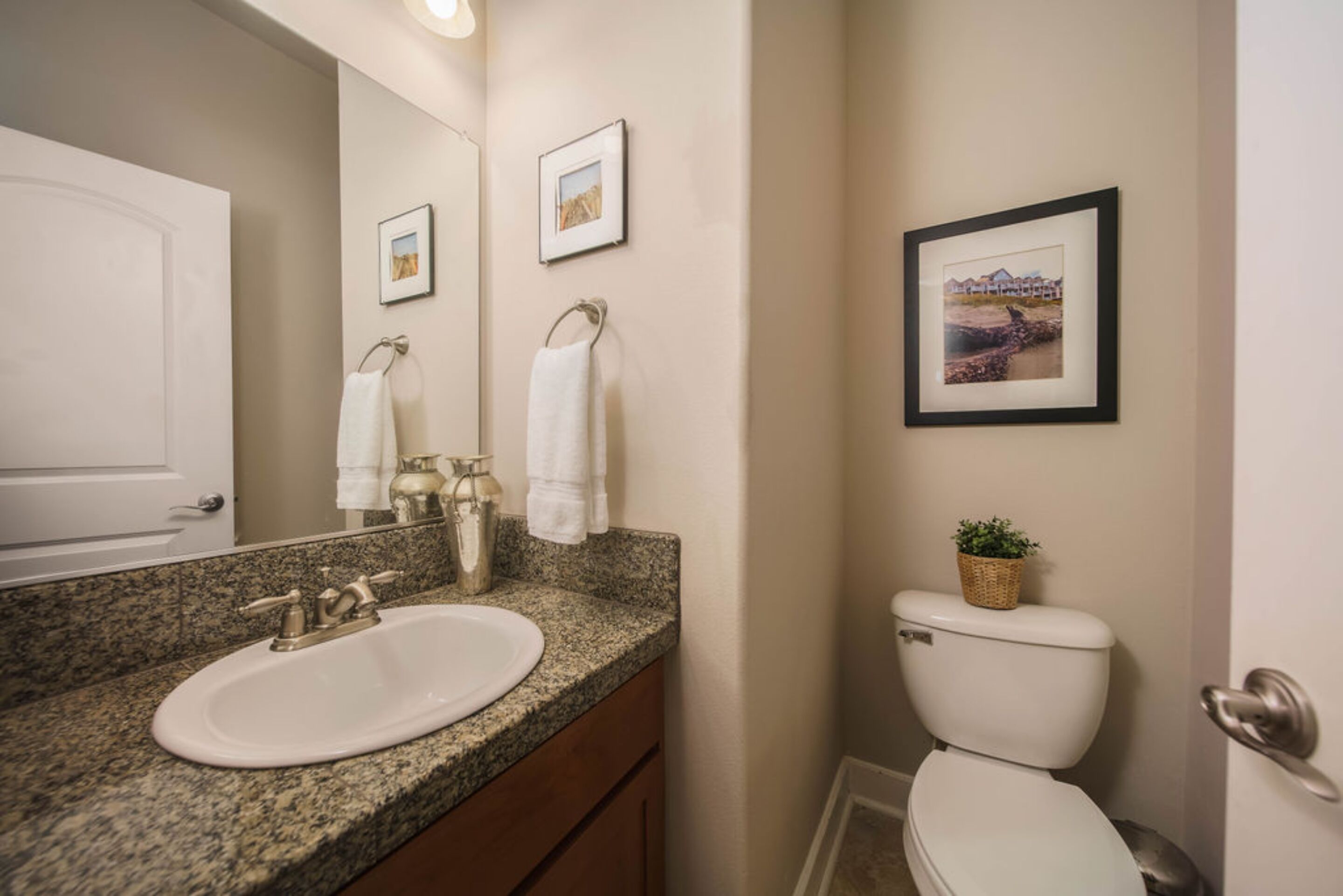A bathroom with a granite countertop sink, a mirror, a toilet, and framed pictures on the wall.
