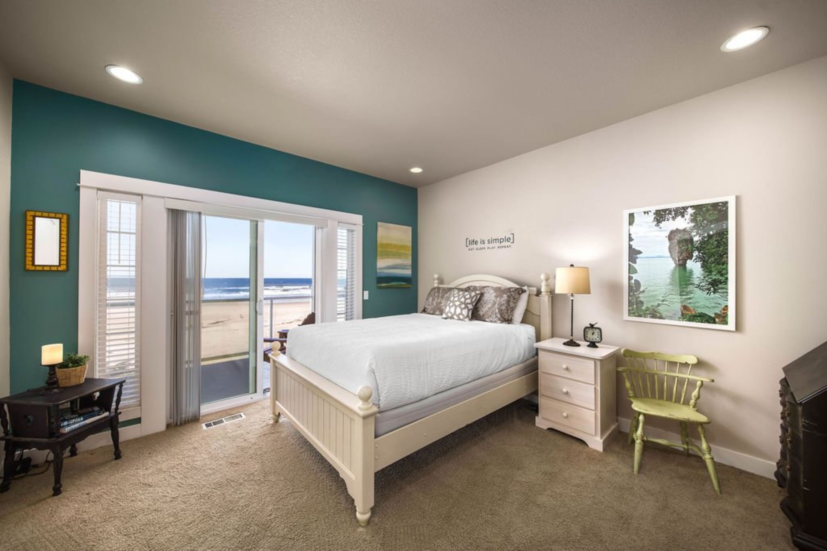 A bedroom with a bed, a nightstand, a chair, a desk, and a view of the beach through a sliding door.