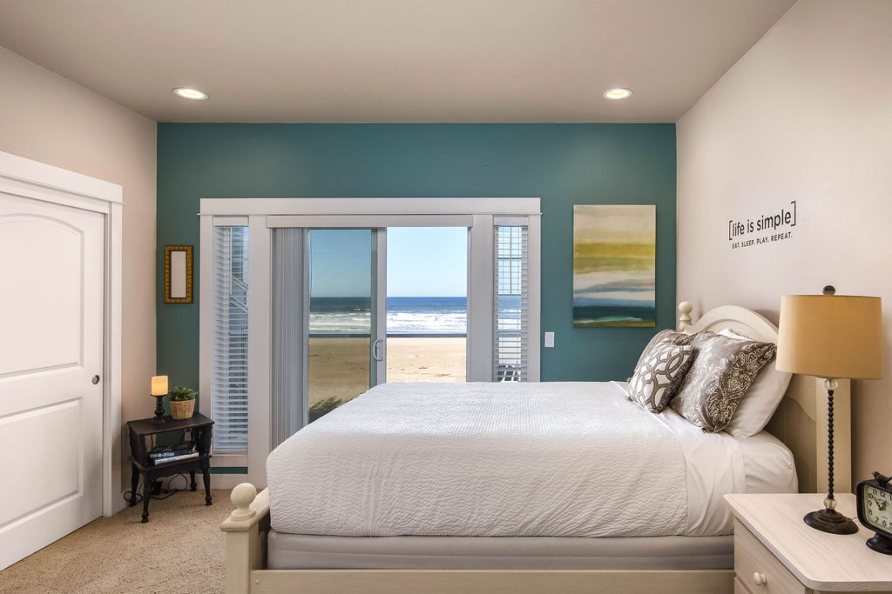 A bedroom with a bed, a nightstand, a lamp, a clock, and a view of the beach through a sliding door.