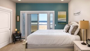 Quarto com cama, mesa de cabeceira, abajur, relógio e vista para a praia através de uma porta deslizante.