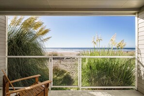 Una silla de madera en un balcón con vista a una playa, hierbas altas y una vista despejada del océano.