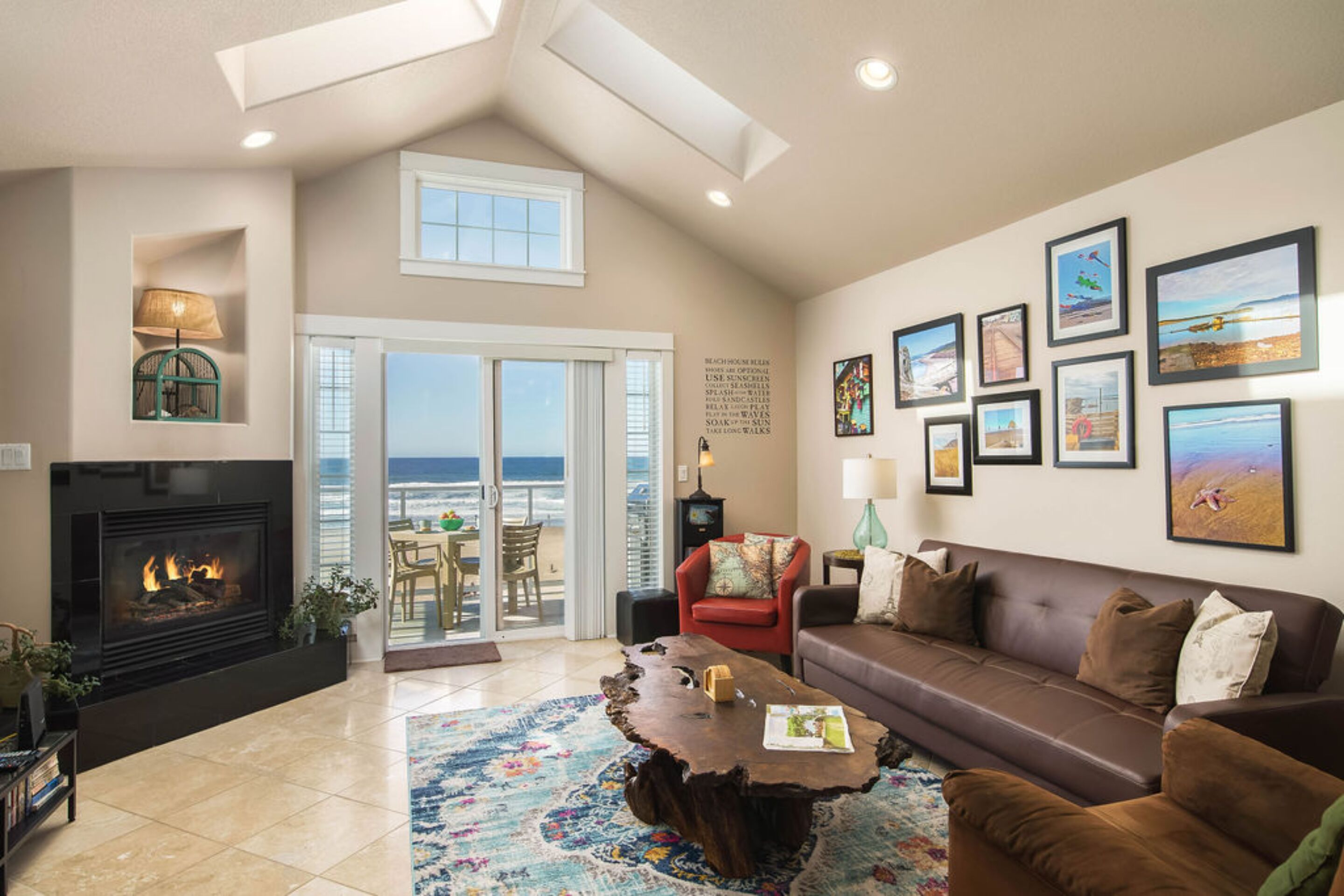 A spacious living room with a fireplace, a large sofa, a coffee table, and a view of the beach through the glass doors.