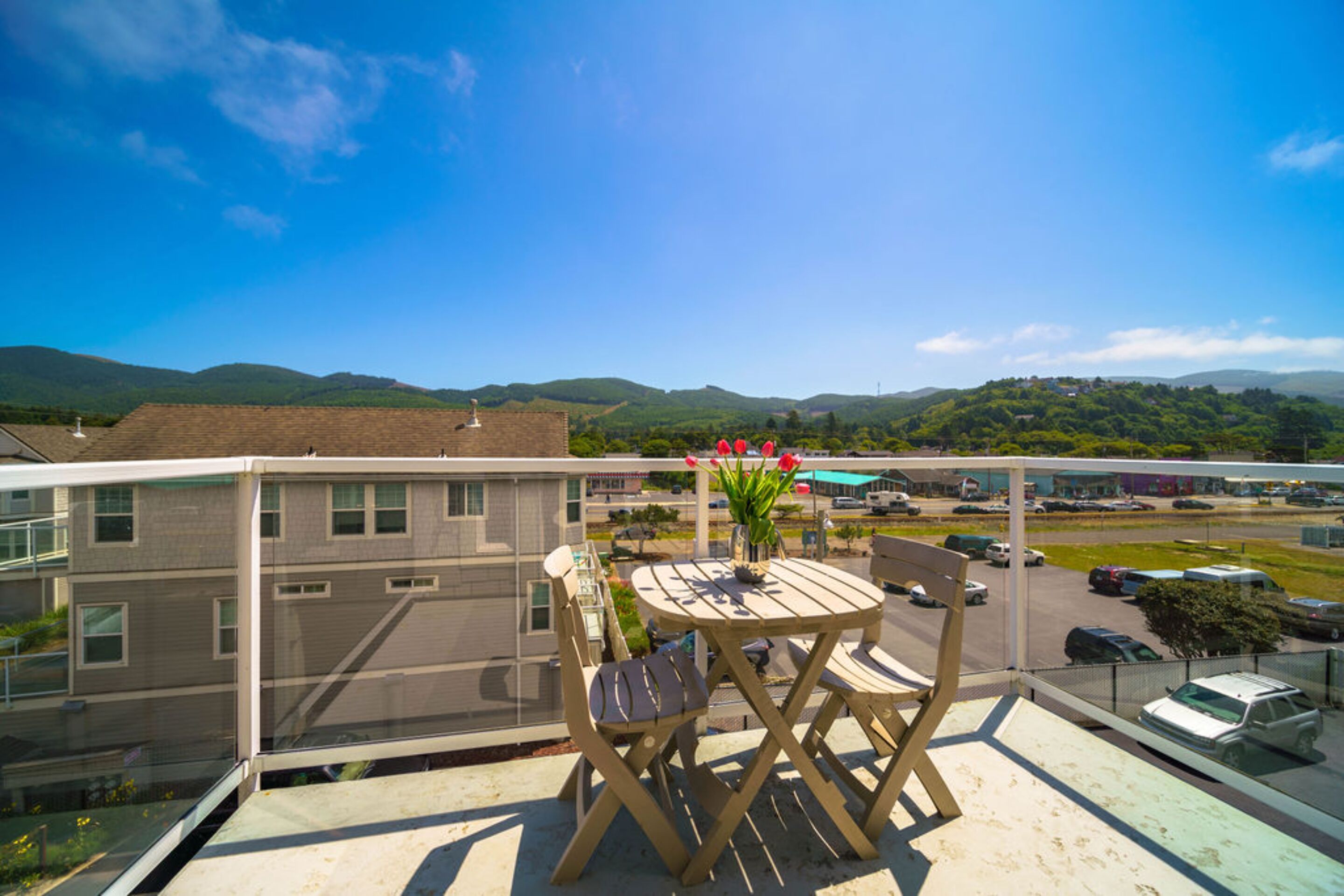 A balcony with a table and chairs, overlooking a parking area and distant hills.