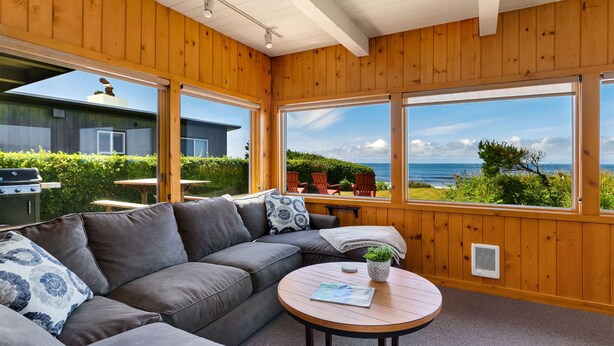 A cozy room with a wooden ceiling, large windows offering a view of the sea, a gray sofa, and a small table with a plant. - The Pilot House by AvantStay | Stunning Home Right On Arch Cape Beach! (Arch Cape)
