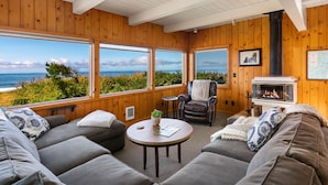 A cozy room with a large window overlooking the ocean, a fireplace, and comfortable seating. - The Pilot House by AvantStay | Stunning Home Right On Arch Cape Beach! (Arch Cape)