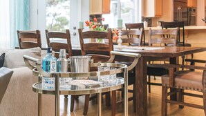 Un coin repas comprenant un chariot en métal sur lequel sont posés une bouteille et des verres, des chaises en bois et une table.