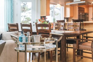 Un coin repas comprenant un chariot en métal sur lequel sont posés une bouteille et des verres, des chaises en bois et une table.