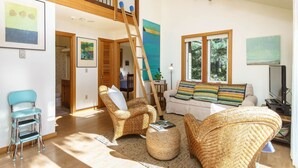Ein geräumiges Wohnzimmer mit hoher Decke, einer Holztreppe, Korbmöbeln und großen Fenstern.