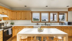Eine moderne Küche mit Holzausstattung, einer großen Kochinsel mit Barhocker, einem eingebauten Backofen und Blick ins Freie durch große Fenster.