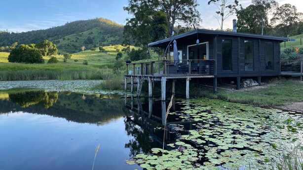 A lakeside cabin with a deck, surrounded by lush greenery and hills.