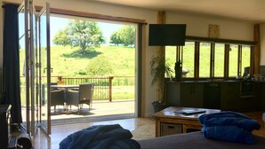 A room with a bed, a table, a chair, a TV, and a view of a green landscape through large windows.