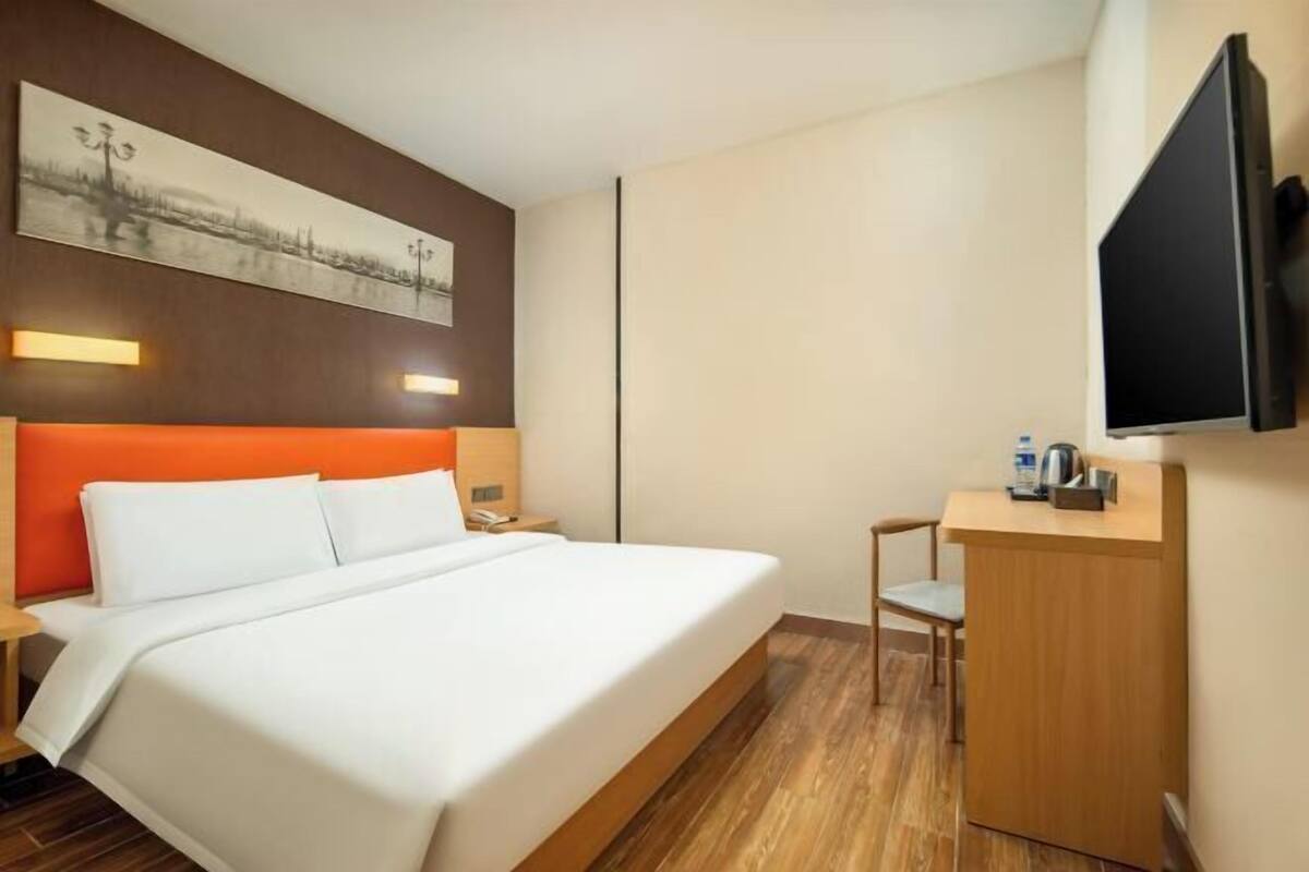 A hotel room with a bed, a desk, a chair, a television, and a painting on the wall.