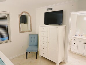 Ein Badezimmer mit weißem Waschtisch, einem großen Spiegel, einem Fernseher über dem Waschtisch und einem blauen Stuhl.