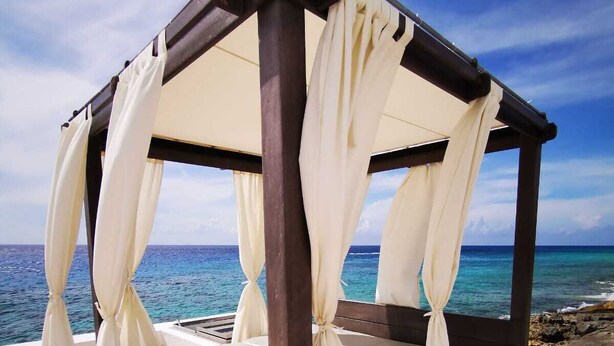 Een bed met hemelbed en witte gordijnen, geplaatst op een balkon met uitzicht op een strand en de oceaan.
