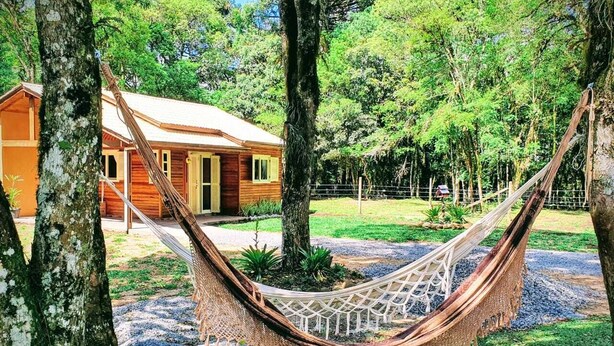 A hammock between trees in a natural setting with a wooden building in the background. - Sitio Recanto Feliz (Gramado)