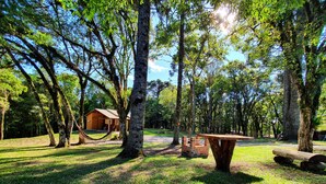 A rustic cabin with a hammock, surrounded by trees and a picnic table in a sunny park setting. - Sitio Recanto Feliz (Gramado)