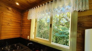 A wooden cabin kitchen with a gas stove, sink, and a window overlooking a forest. - Sitio Recanto Feliz (Gramado)