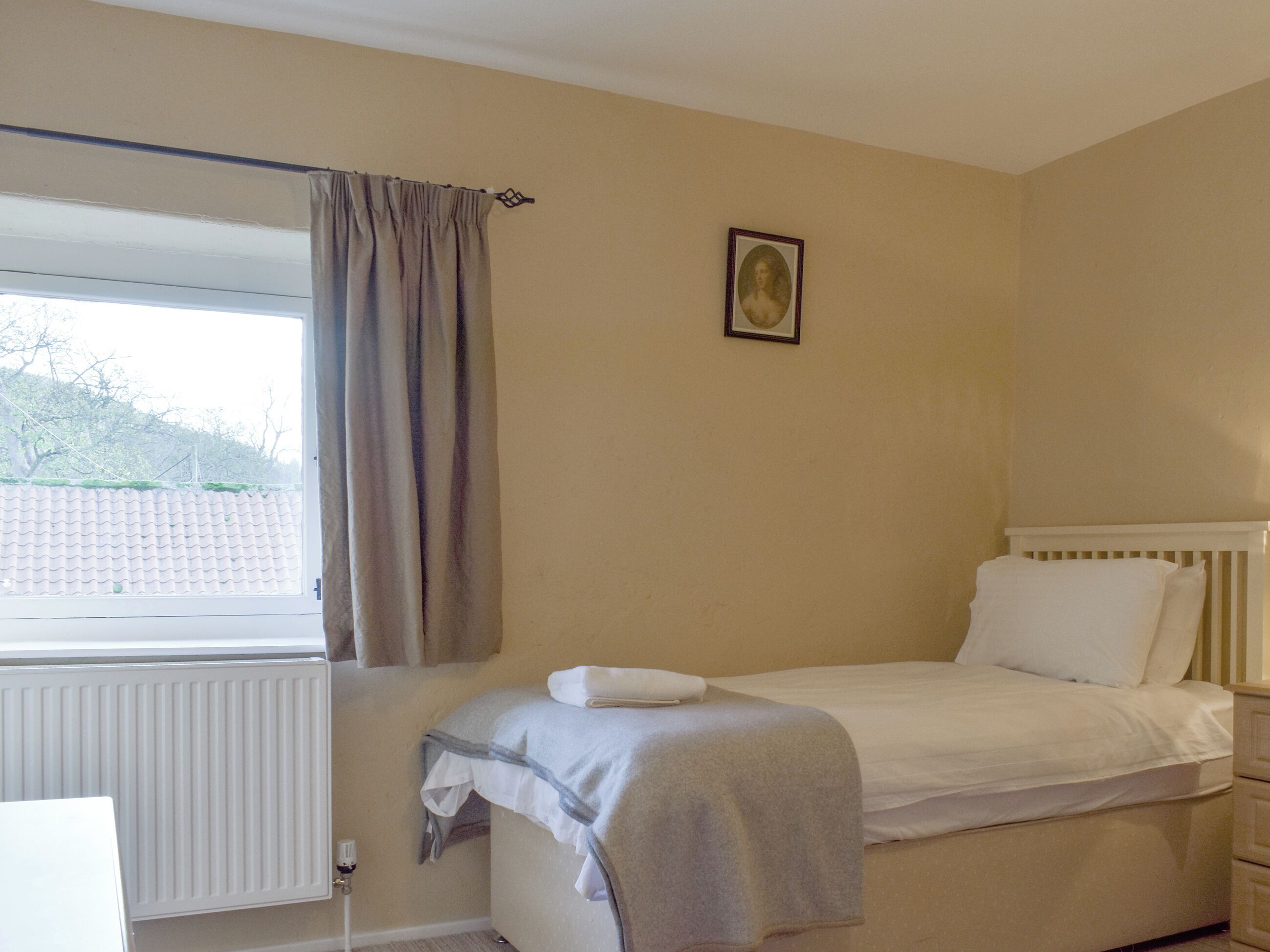 A bedroom with a single bed, a window with curtains, a radiator, and a framed picture on the wall.