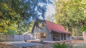 Eine rustikale Holzhütte mit rotem Dach, einer Laube mit Lichterketten und einem Kieshof, umgeben von Bäumen.