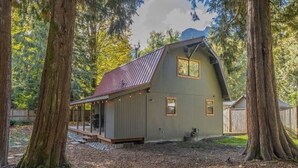 Eine rustikale Holzhütte mit geneigtem Dach, umgeben von hohen Bäumen und einer Holzterrasse.