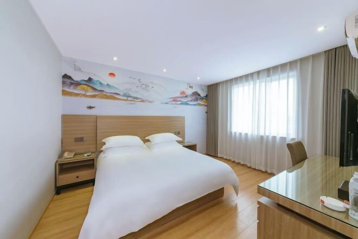 A hotel room with a bed, a desk, a chair, a nightstand, and a wall with a painting.