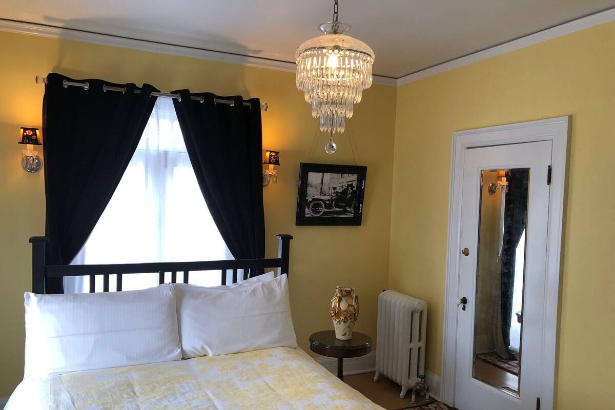 A bedroom with a bed, black curtains, a chandelier, a radiator, a door, and a framed picture on the wall.