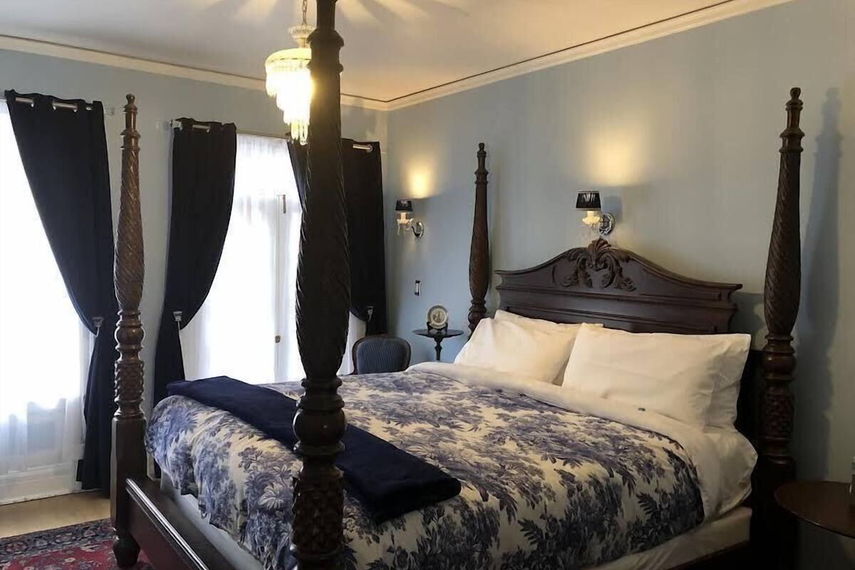 A four-poster bed with a floral bedspread, a bedside table with a lamp, and a view of the outdoors through a window with curtains.