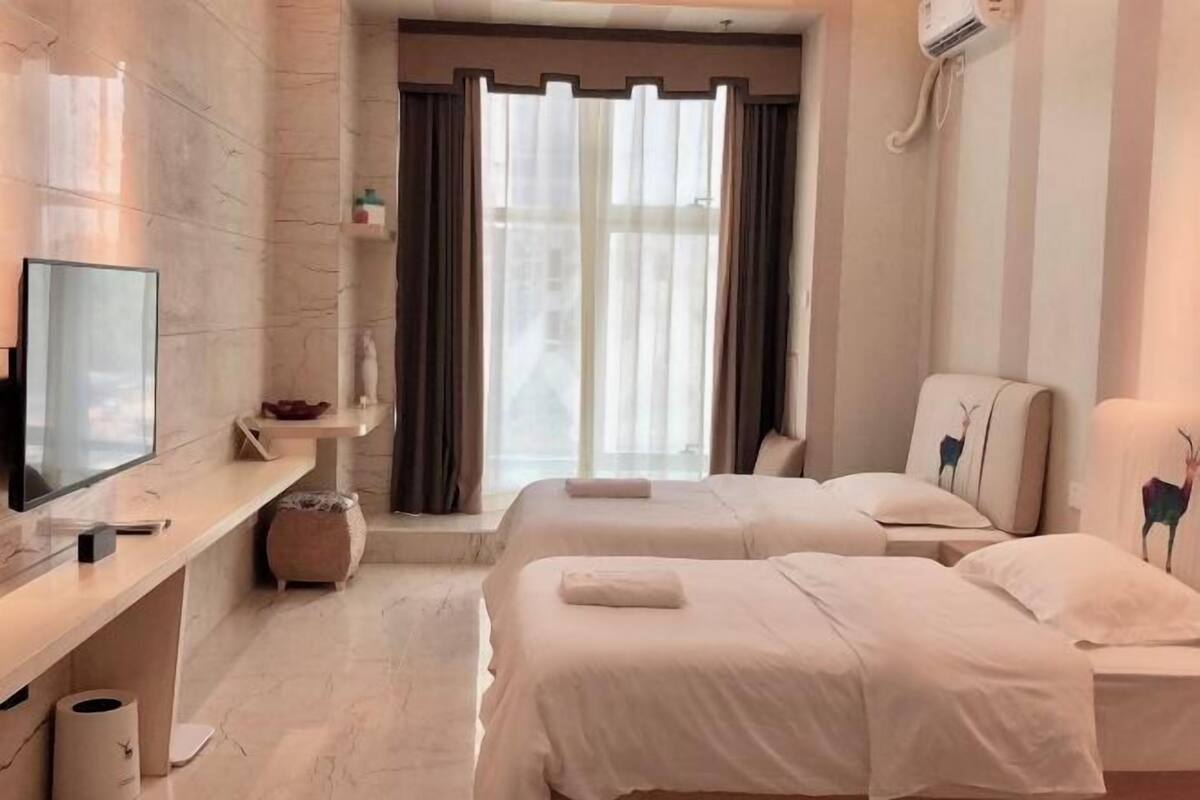 Hotellihuone, jossa on kaksi sänkyä, televisio, pieni pöytä ja ikkuna, jossa on verhot.