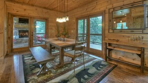 Ein Holzhütten-Interieur mit Kamin aus Stein, einem Holzesstisch, Stühlen, einer Bank, einem Teppich und großen Fenstern.