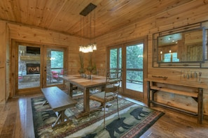 Ein Holzhütten-Interieur mit Kamin aus Stein, einem Holzesstisch, Stühlen, einer Bank, einem Teppich und großen Fenstern.