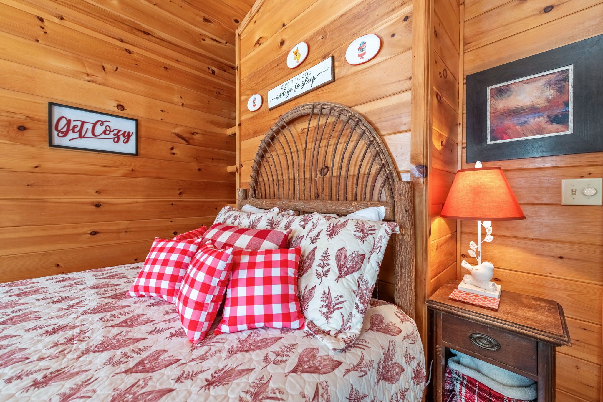 A cozy wooden room with a bed, checkered pillows, a nightstand with a lamp, and wall decorations including a 'Get Cozy' sign and a 'Give it to God will go to sleep' sign.