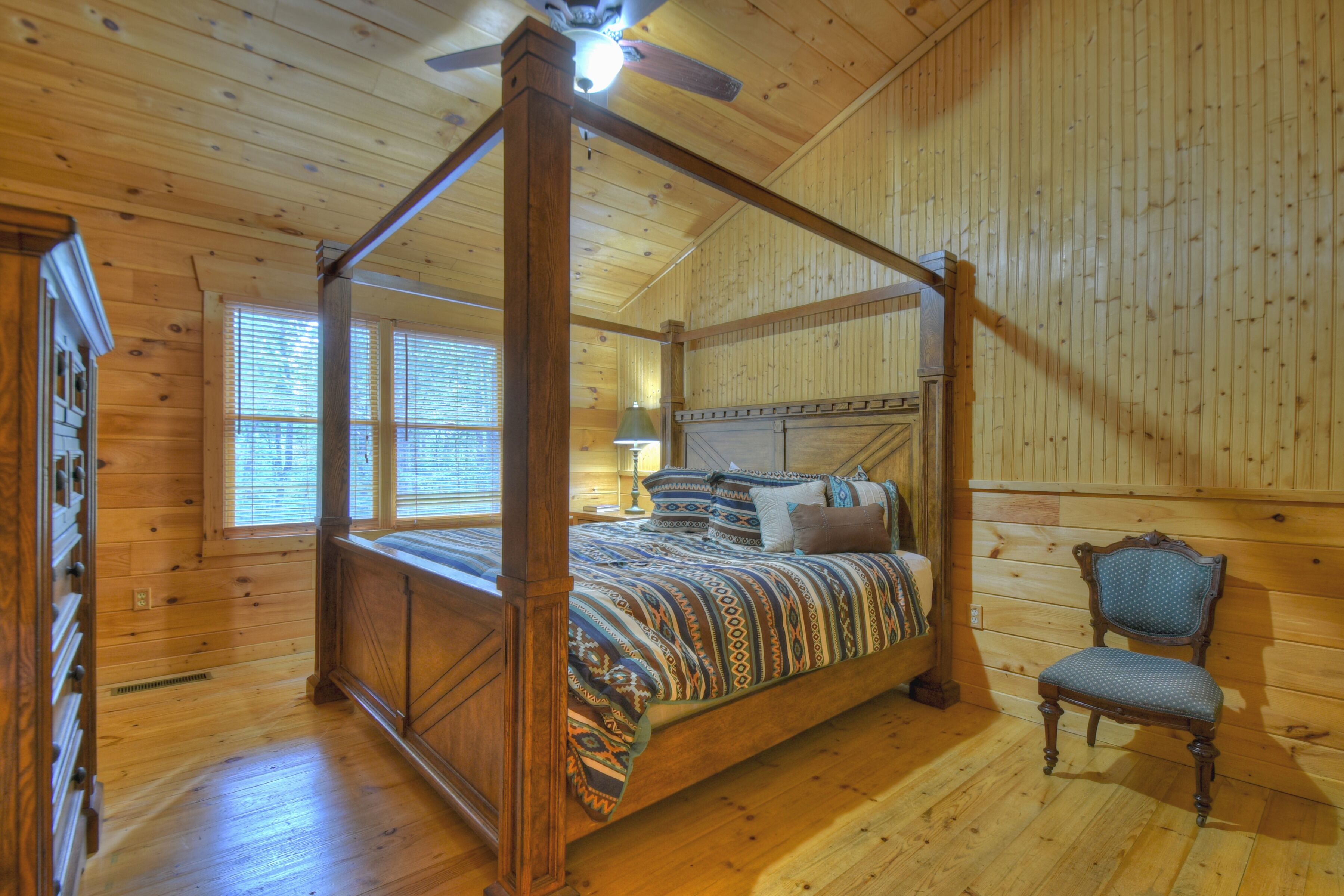 A four-poster bed with a patterned bedspread, a wooden chair, and a window with blinds.