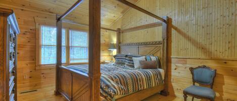 A four-poster bed with a patterned bedspread, a wooden chair, and a window with blinds.